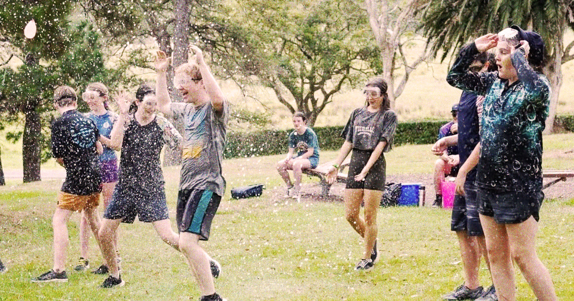 Eight young people laughing and playing in a park as water is sprayed in the air.