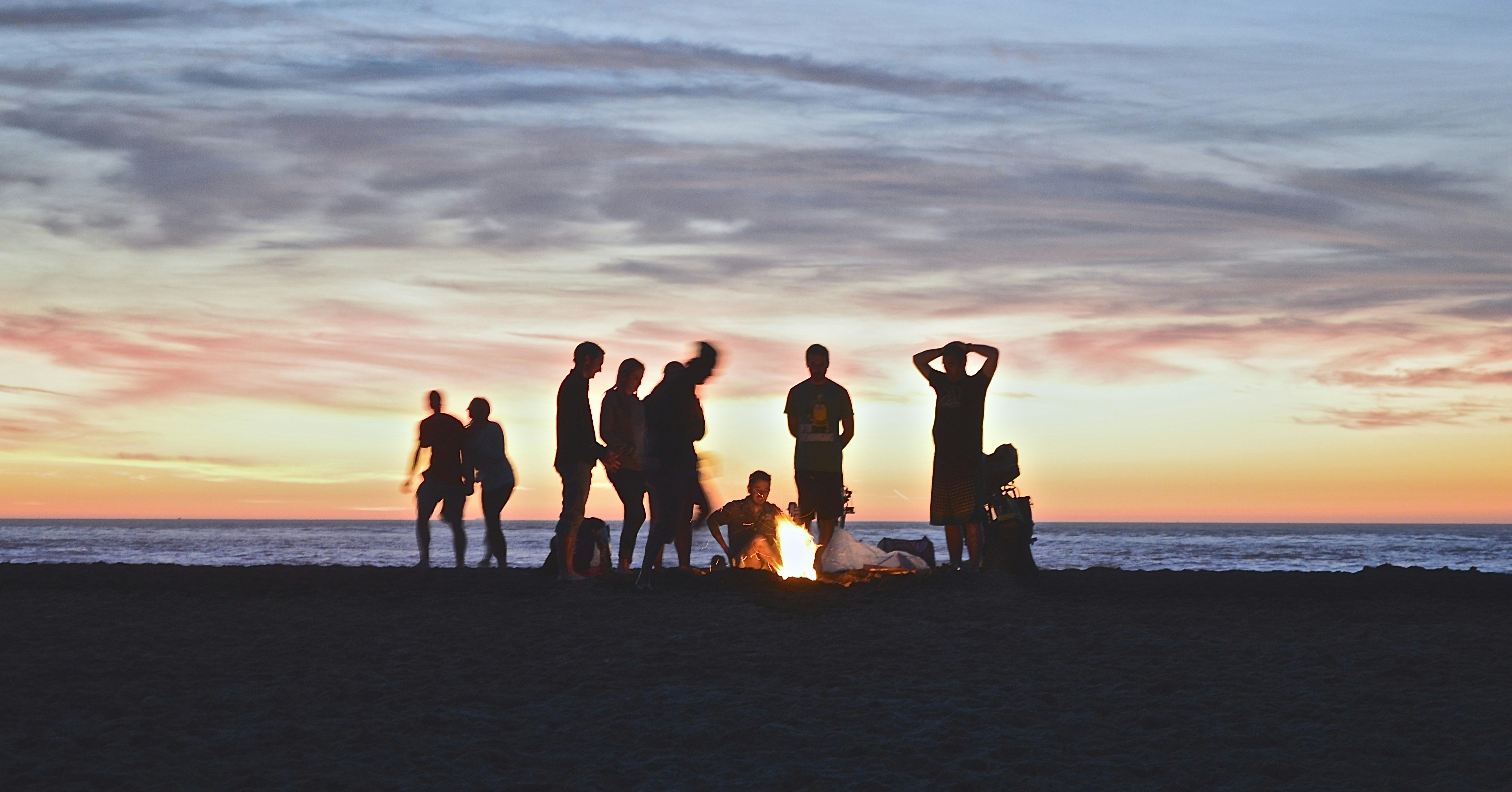 A silhouette of a small group of young people around a campfire at a beach during sunset.